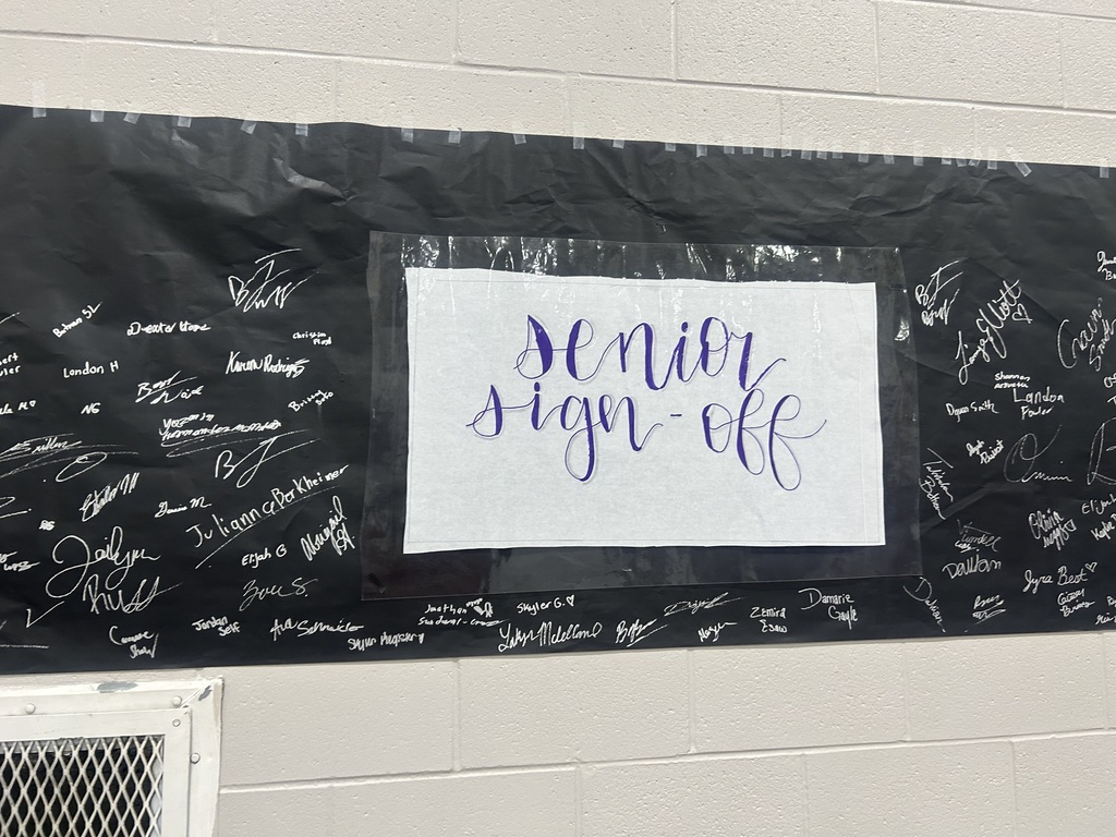 A black wall display filled with student signatures surrounds a sign that reads “Senior Sign Off” in purple lettering.