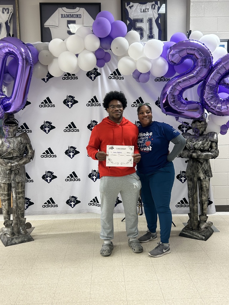 A student and an adult stand together in front of a school Decision Day backdrop with purple and white balloons and “2026” balloon numbers. The student holds an “I Decided!” certificate while both smile at the camera, with knight statues on either side.