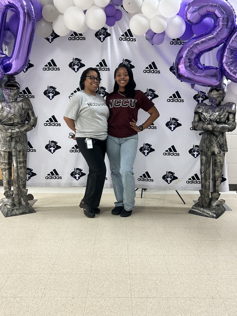 Two people stand side by side smiling in front of a school-branded backdrop with purple and white balloons and knight statues. One wears an NCCU shirt, showing school pride during Decision Day.