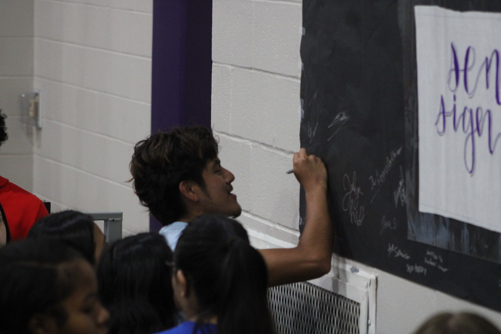 A student signs their name on a black wall or board filled with other signatures while classmates stand nearby watching. A sign partially visible on the wall reads “Senior Sign Off.”