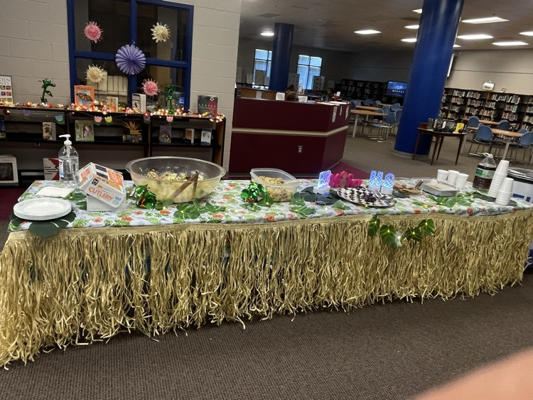 A library with tables lined with grass skirts on the edges, sign in the middle of the table that says aloha and bowls of food for teacher appreciation week