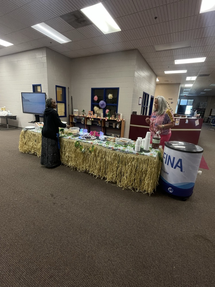 A library with tables lined with grass skirts on the edges, sign in the middle of the table that says aloha and bowls of food for teacher appreciation week 