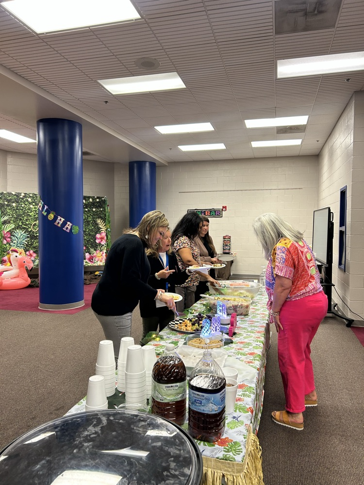 A library with tables lined with grass skirts on the edges, sign in the middle of the table that says aloha and bowls of food for teacher appreciation week 