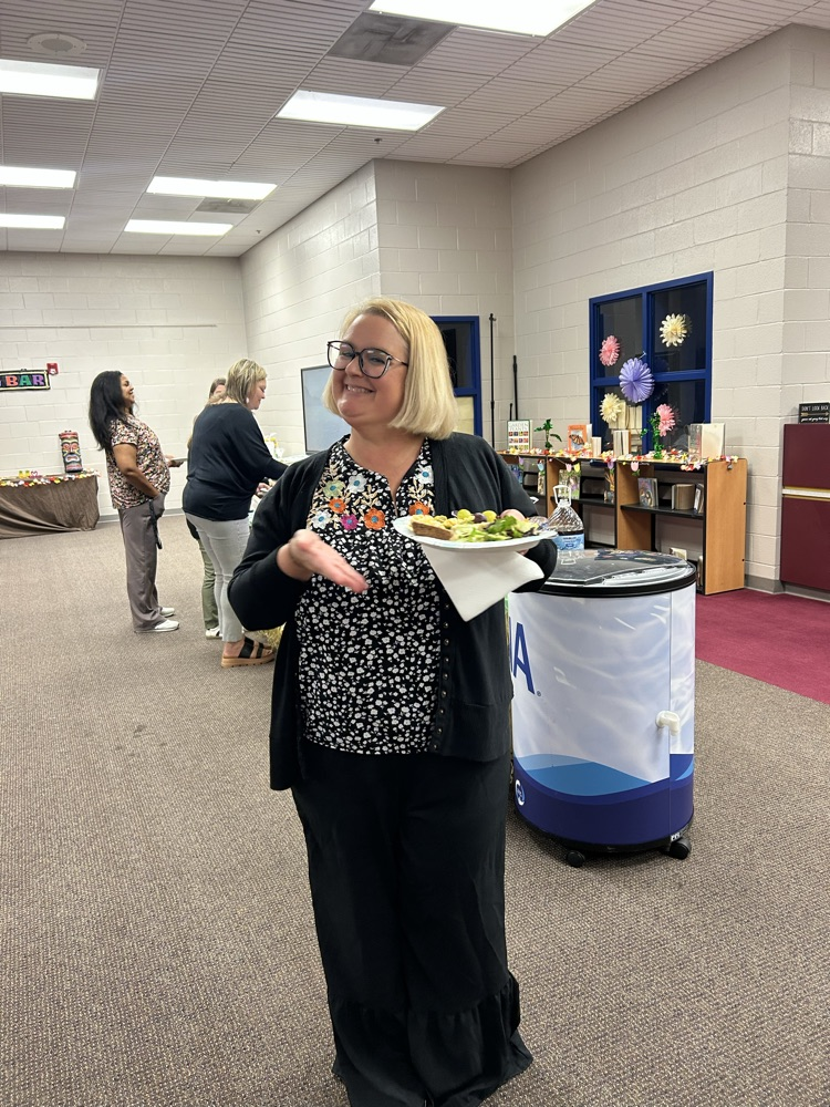 A lady with blonde hair, a black skirt and black and white top shows off her plate of food that she has fixed for teacher appreciation
