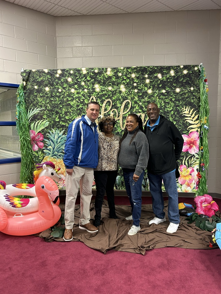 Four people staying in front of a aloha Hawaiian backdrop there is a unicorn and flamingo pool floaty to the left. The people are smiling as they stay in front of the backdrop and Hawaiian decorations.