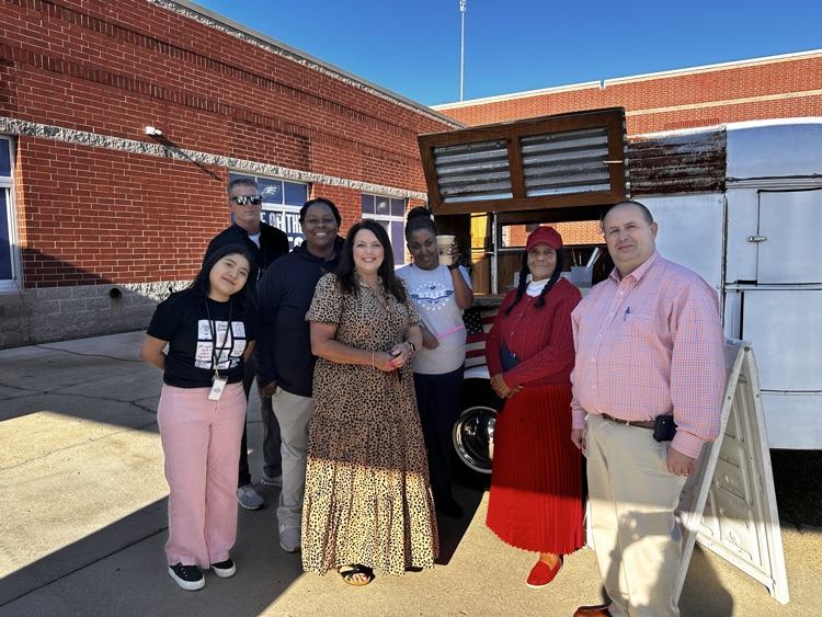 A group of staff members stand together smiling outside. behind them is a coffee cart.
