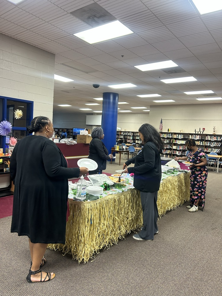 three teachers fix a plate of food by a Hawaiian themed table with a grass skirt