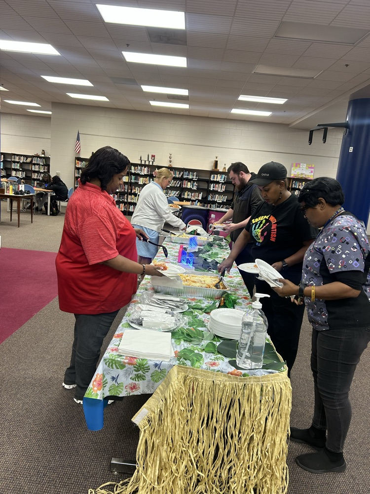 three teachers fix a plate of food by a Hawaiian themed table with a grass skirt
