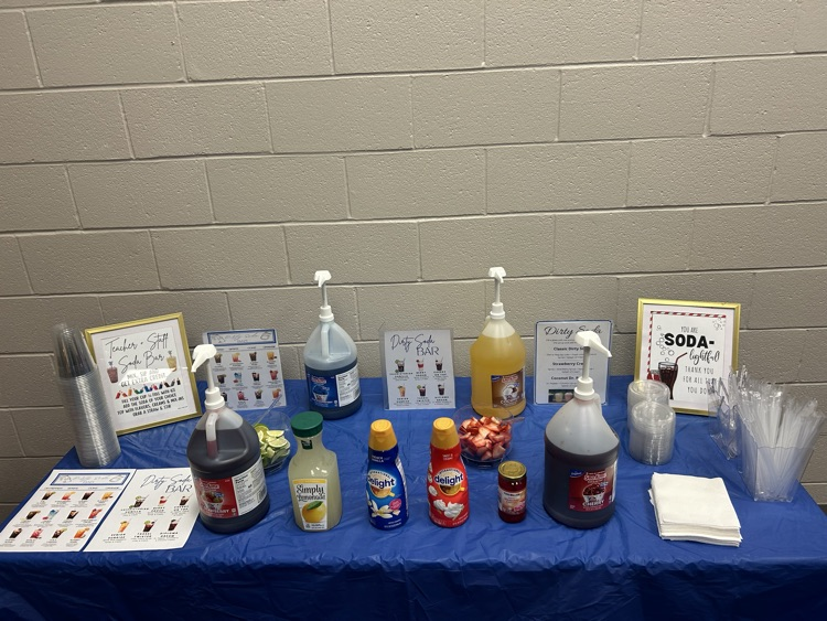 Table with blue table cloth, gallons of syrups and soads, and menus for a dirty soda bar