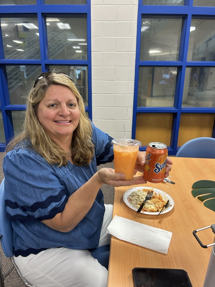 teacher in blue shirt and white pants with a cup of orange drink and a sunkist can. there is a plate of food on the table
