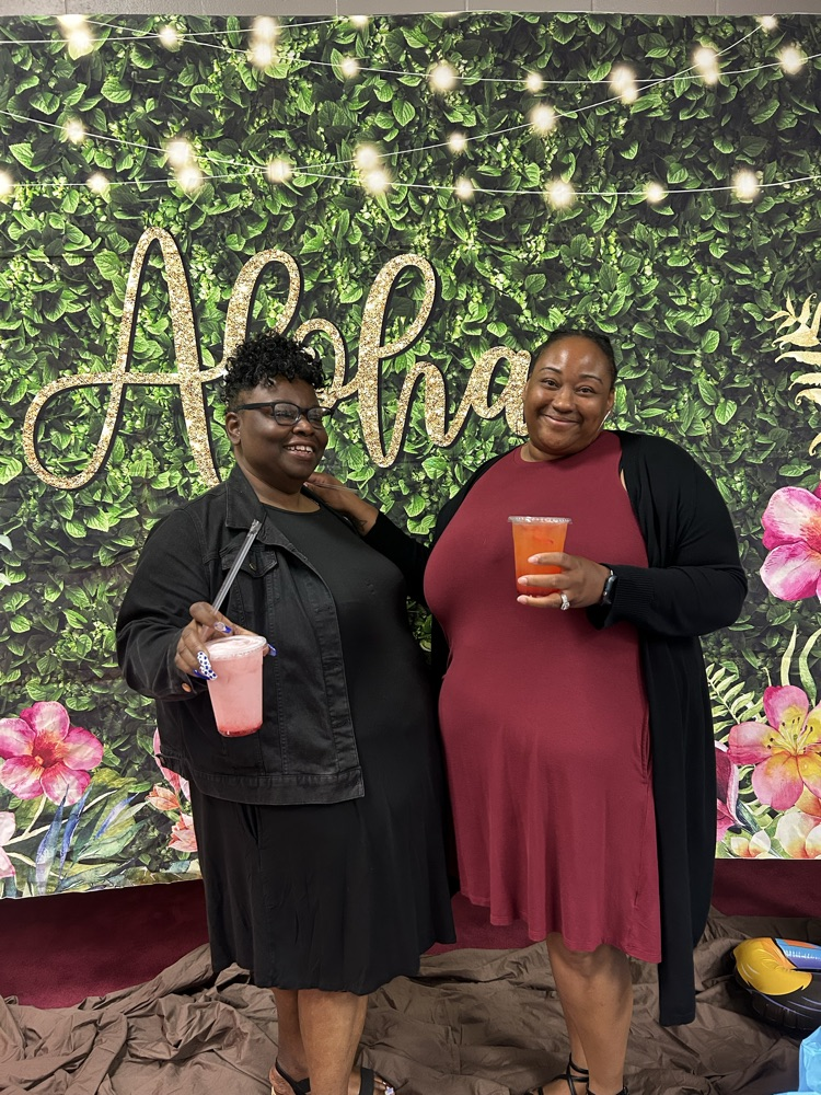 teacher in black dress and teacher in burgundy dress in front of a aloha Hawaiian backdrop with their refreshing drinks