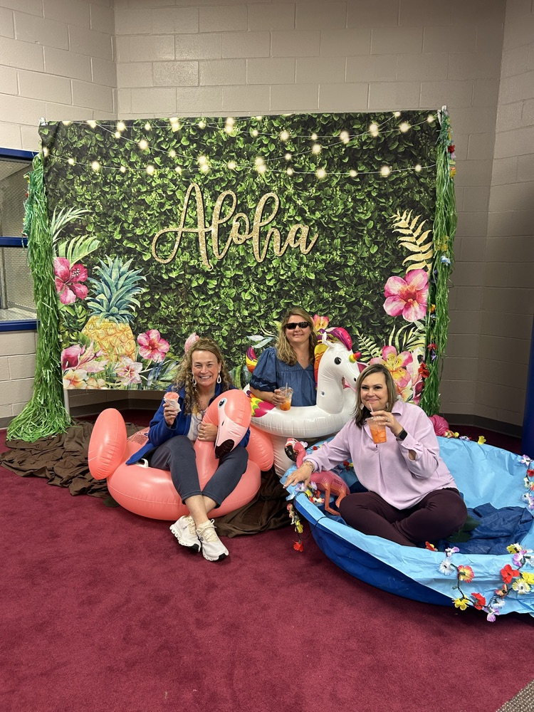 teacher on a flamigo float, teacher with a unicorn float, and a teacher in a kiddy pool posing with their dirty sodas in front of the aloha Hawaiian backdrop