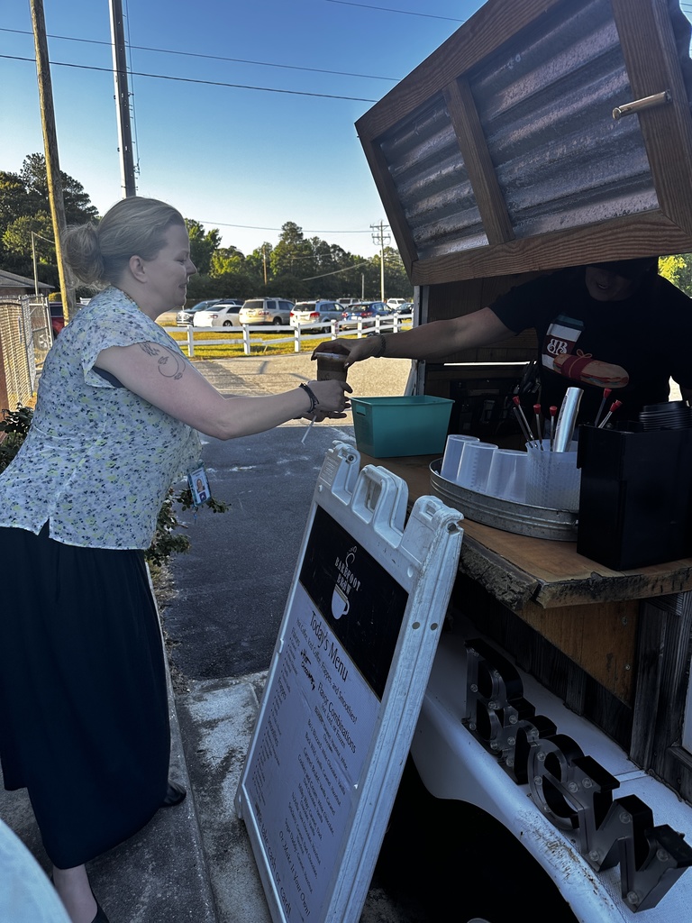 A woman with a school ID badge reaches across to receive a drink from a worker inside the Barefoot Brew mobile coffee trailer, which displays a handwritten menu board outside.