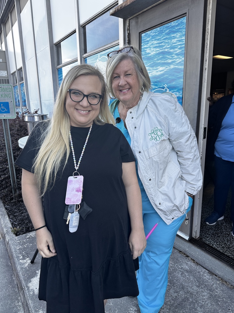 Two women smile together outside a school entrance with ocean-themed window graphics. One wears a black dress with a beaded lanyard, the other is in teal scrubs and a white monogrammed jacket.