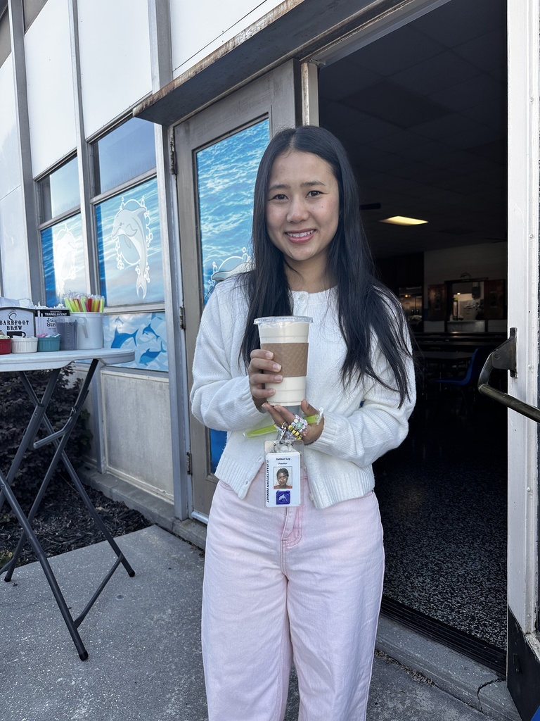 A young woman wearing a white sweater and pink pants holds an iced drink and smiles outside a school entrance. She wears a school ID badge on a beaded lanyard. A Barefoot Brew setup is visible in the background.