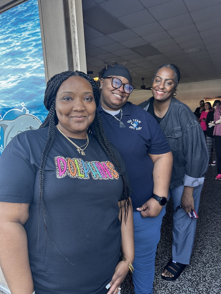Three women smile for the camera inside a school hallway. The woman in front wears a black "Dolphins" school spirit shirt, and a dolphin-themed mural is visible on the window beside them.