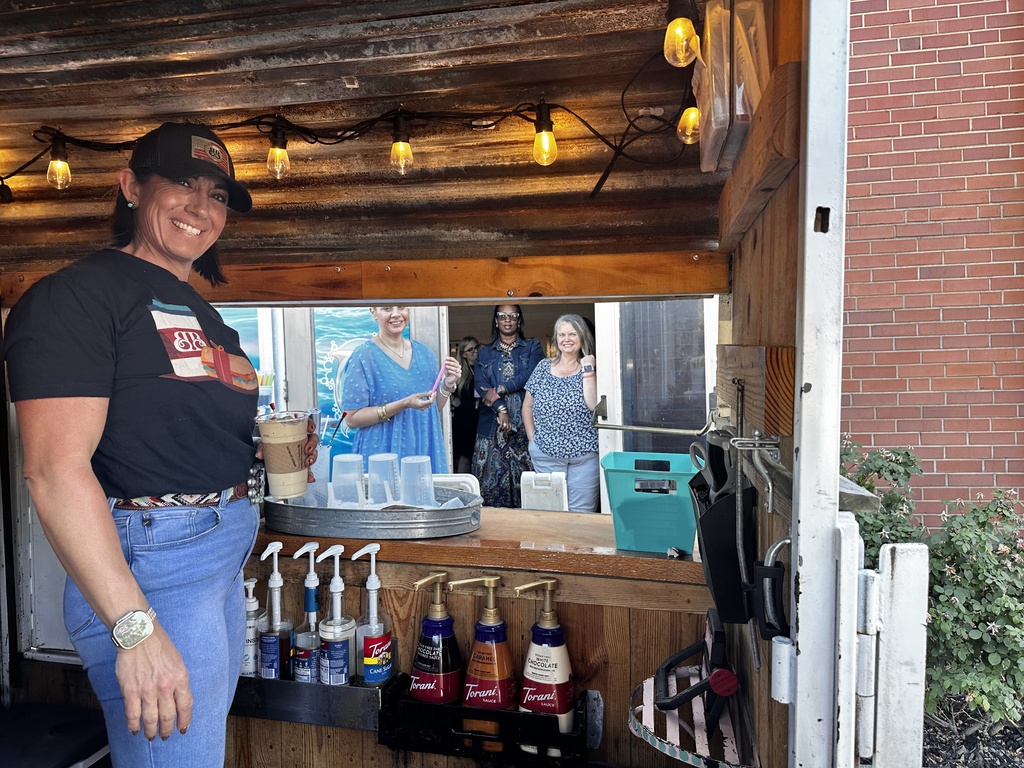 A woman in a black Barefoot Brew T-shirt and baseball cap smiles beside the service window of a rustic wooden coffee trailer. Several teachers wait in line through the window behind her, and Torani syrup bottles are visible on the counter.