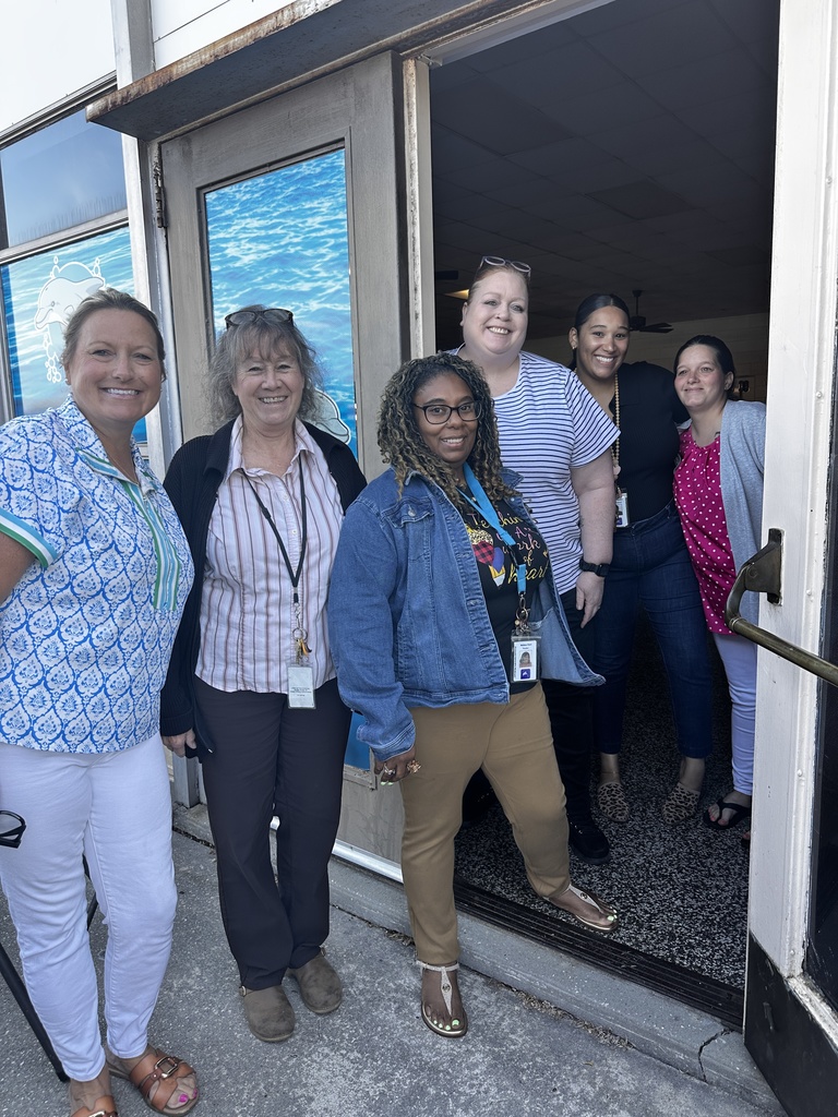 Five school staff members pose together smiling in and around a school entrance with ocean-themed door graphics. Several wear school ID badges and lanyards.