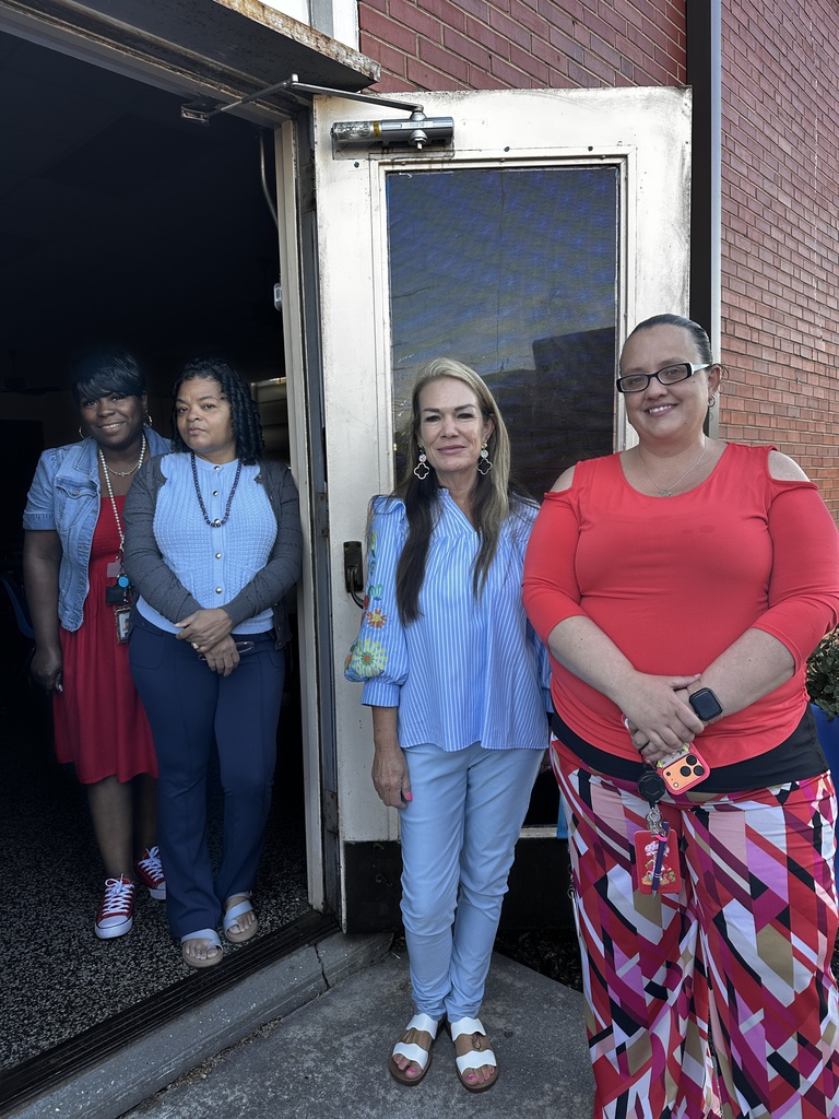 Four women stand together outside a brick building near a door, smiling at the camera. They are dressed in colorful attire including red, blue, and patterned outfits, with some wearing school ID badges.