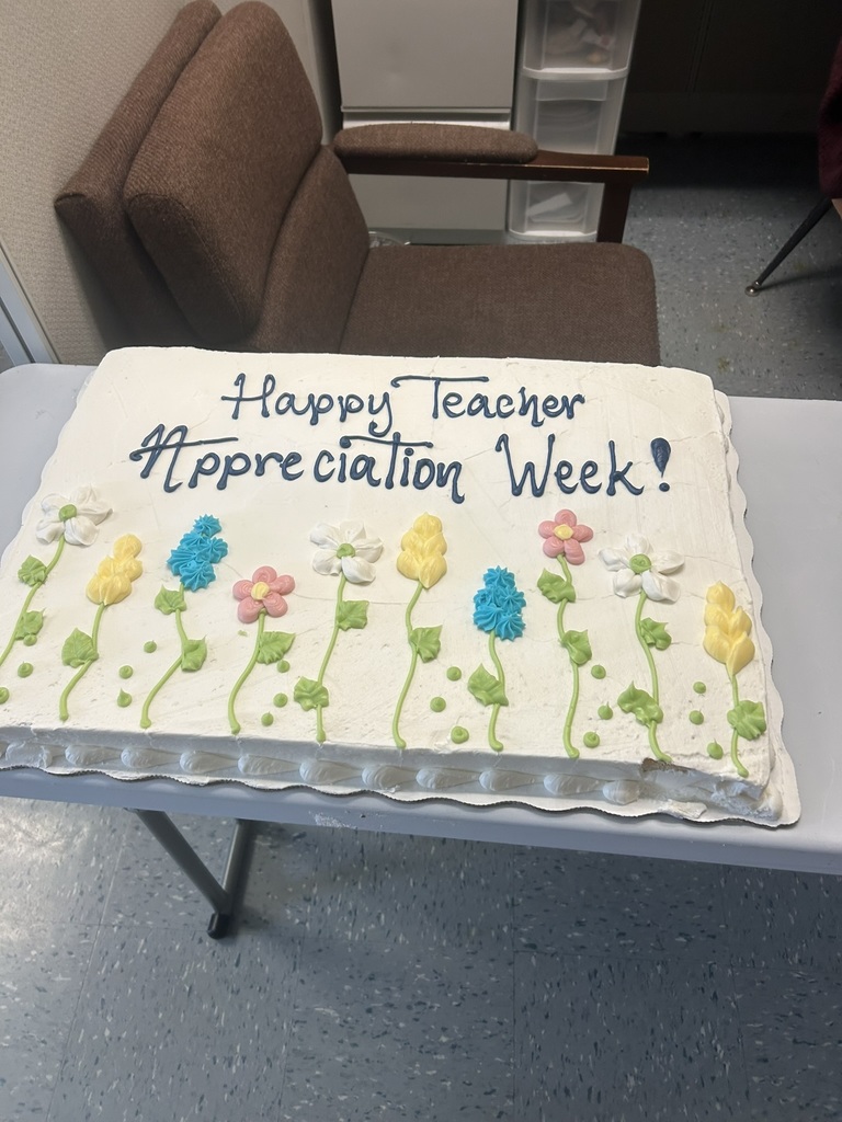 A large sheet cake decorated with colorful icing flowers sits on a table. The cake reads “Happy Teacher Appreciation Week!” in blue icing.