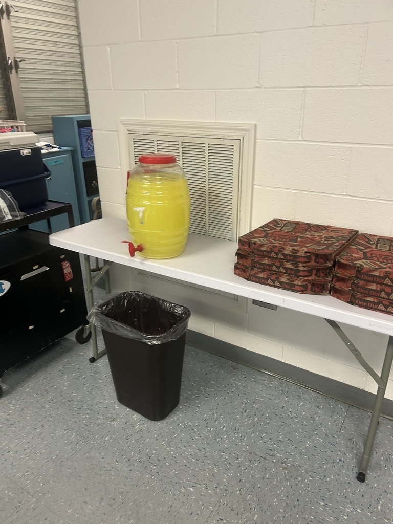 A large container of yellow drink and stacks of pizza boxes are set up on a table as part of a Teacher Appreciation Week lunch spread.