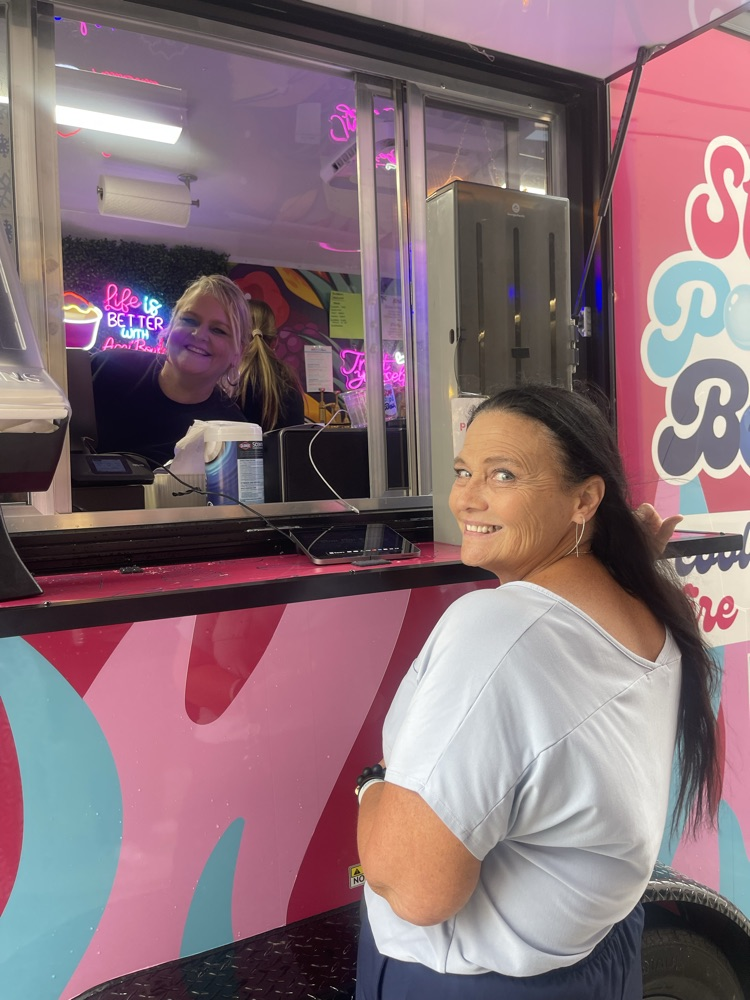 A woman inside of a food truck, smiling while a woman waits for her food, smiling at the camera