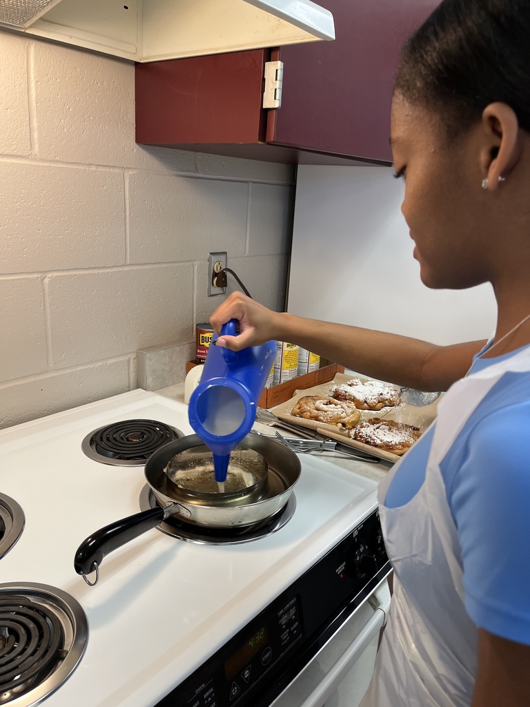 A student in a white apron stands at a kitchen counter, focusing on preparing food in a frying pan.