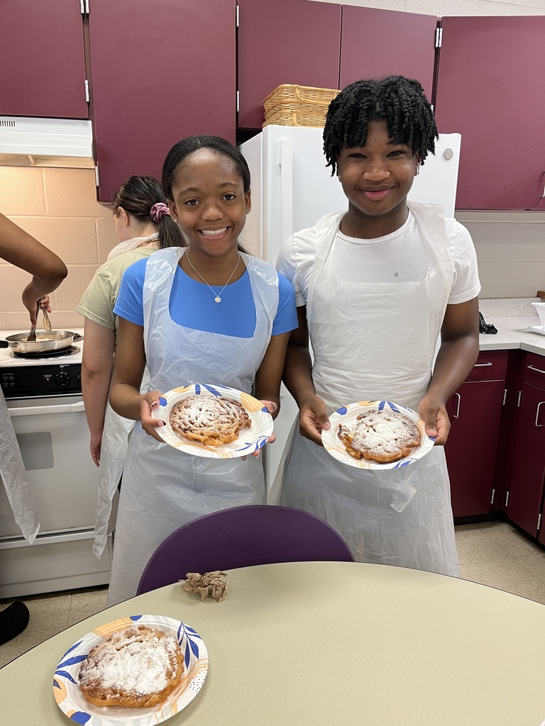 Two students standing in a kitchen, proudly holding up plates of food they have cooked, with a classroom setting in the background