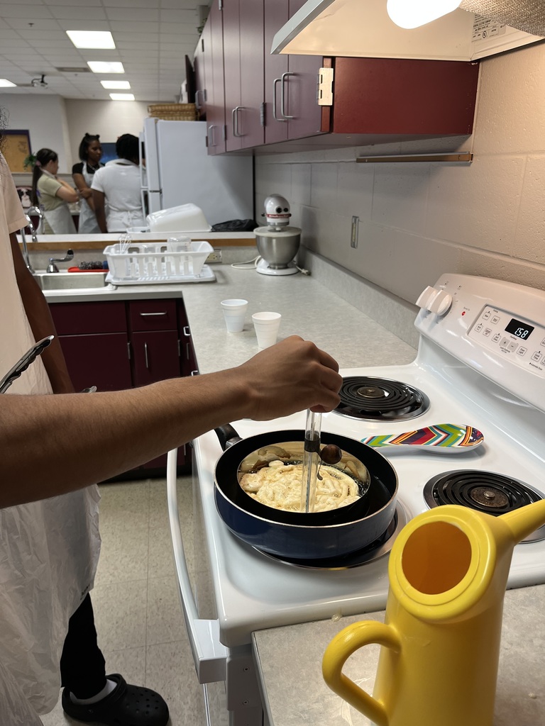 A close-up view of a person using a spatula to flip food in a frying pan on a white stovetop in a classroom kitchen