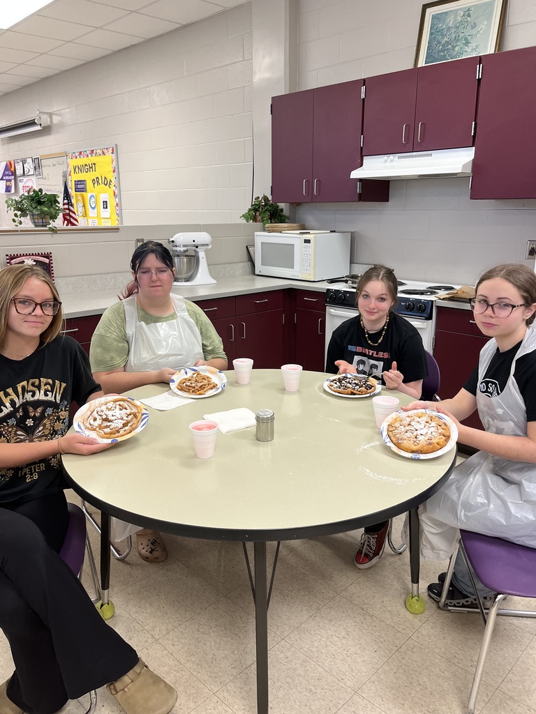 our female students wearing aprons sit at a round table in a school kitchen classroom, smiling and displaying plates of food they have prepared