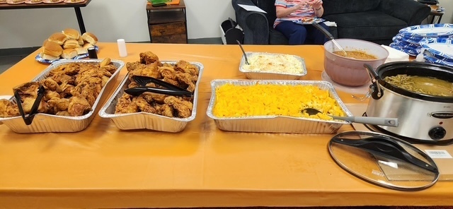 A Teacher Appreciation Week meal spread featuring fried chicken, macaroni and cheese, rolls, baked beans, mashed potatoes, and green beans arranged on an orange-covered table for ETMS staff.
