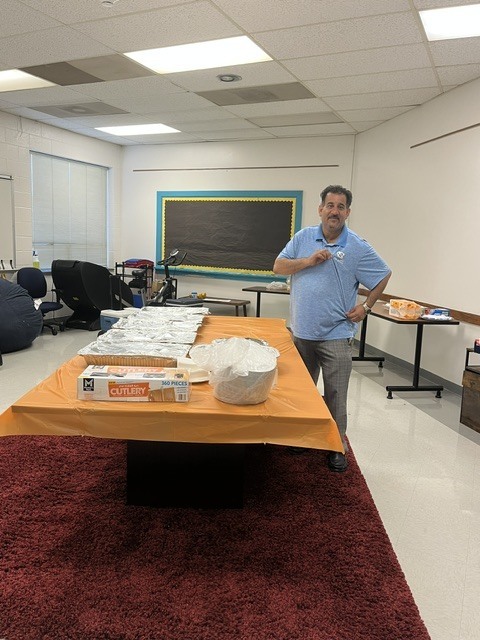 A community supporter stands beside a table filled with covered meal trays, utensils, and serving supplies prepared for ETMS Teacher Appreciation Week celebrations.