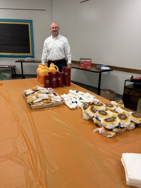 A community supporter stands behind a breakfast spread with biscuits, sandwiches, juice, and coffee supplies arranged on an orange-covered table for ETMS staff appreciation.