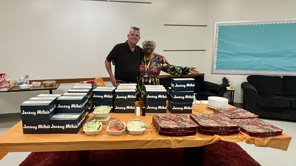 Two community members stand behind a large Teacher Appreciation Week meal setup featuring stacks of Jersey Mike’s sandwich boxes, pizza boxes, salad toppings, plates, and drinks in a decorated ETMS staff room.
