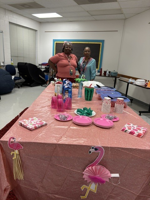 Two staff members stand behind a pink flamingo-themed table display with drinks, snacks, cups, plates, and decorations prepared for Teacher Appreciation Week festivities at ETMS.