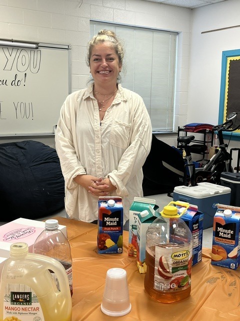 A smiling staff member stands behind a Teacher Appreciation Week refreshment table featuring juices, tea, drinks, cups, and sweet treats prepared for ETMS staff.