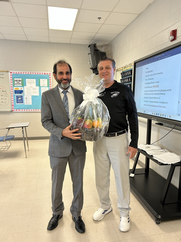 Mr. Deswal stands next to Dr. Carroll in a classroom as he is presented with a fruit basket