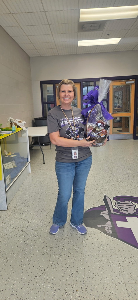 A smiling staff member at West Bladen High School holds a large gift basket wrapped in purple cellophane with a matching bow. She is standing in a school hallway near a doorway.