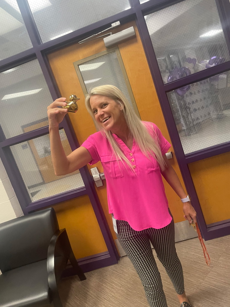A smiling staff member at West Bladen High School holds a large gift basket wrapped in purple cellophane with a matching bow. She is standing in a school hallway near a doorway.