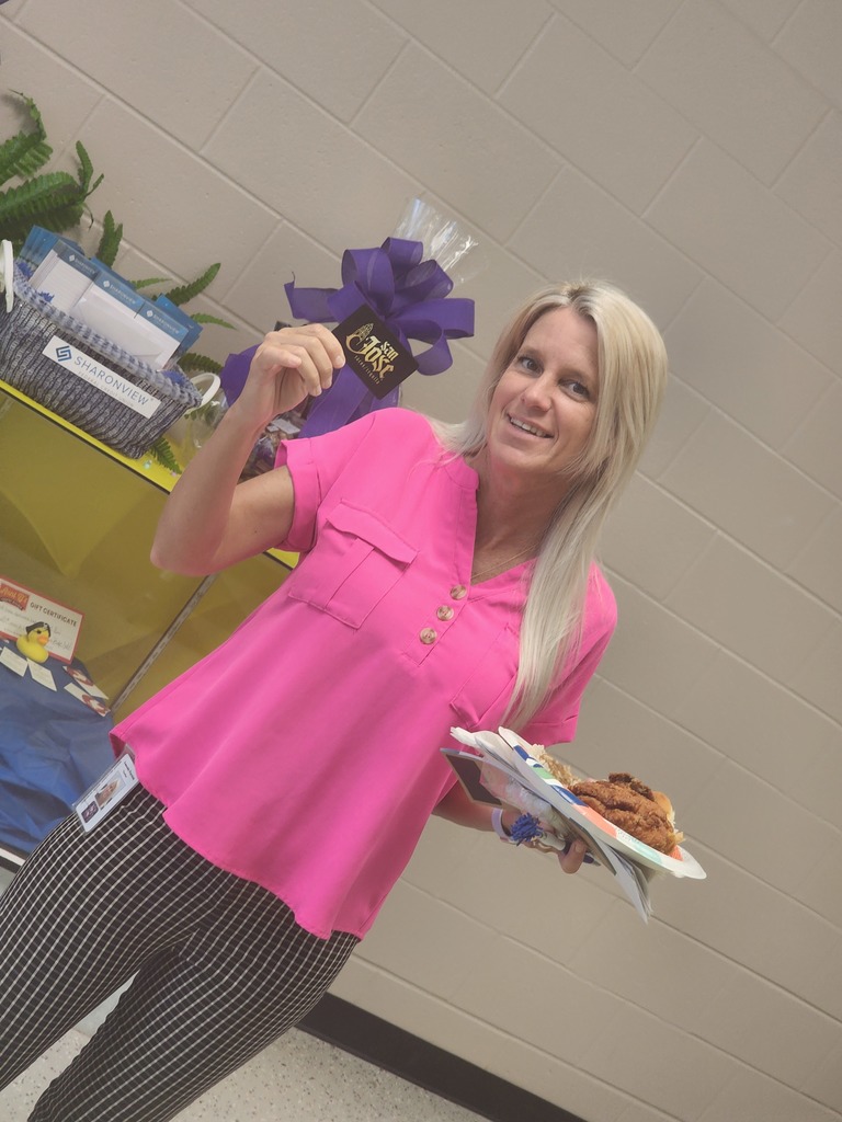 Four women stand together in a school office holding a framed "Teacher Appreciation" certificate and gift bags. They are smiling in front of a purple wall featuring the West Bladen High School "WB" logo.