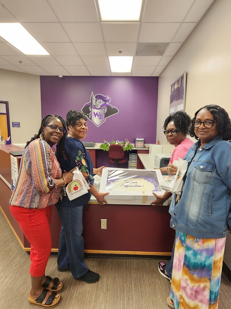 Four women stand together in a school office holding a framed "Teacher Appreciation" certificate and gift bags. They are smiling in front of a purple wall featuring the West Bladen High School "WB" logo.