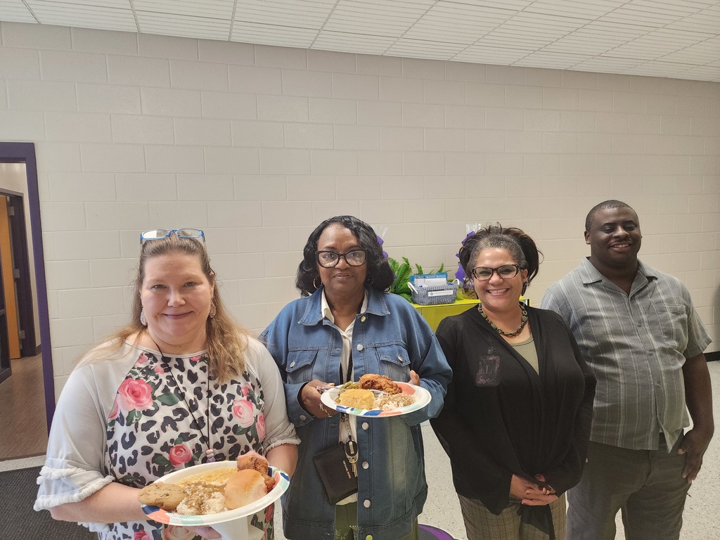 Four staff members at West Bladen High School smile while holding plates of food, including fried chicken and sides, during a staff appreciation luncheon.