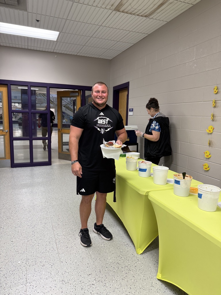 A man in a black West Bladen Football t-shirt smiles while holding a plate of food at a catered staff event. He stands next to a yellow-clothed table featuring various toppings and condiments.