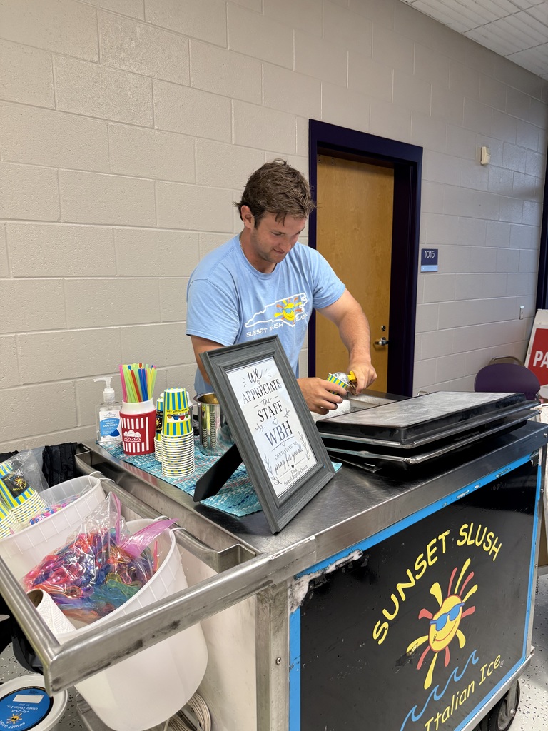A man in a blue t-shirt prepares treats at a Sunset Slush Italian Ice cart. A framed sign on the cart reads, "We appreciate our staff! WBH