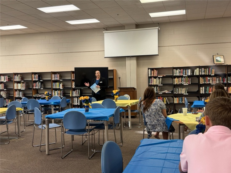 Library with tables that have blue and yellow table clothes with small floral arrangements. a lady is standing at the front talking to the group