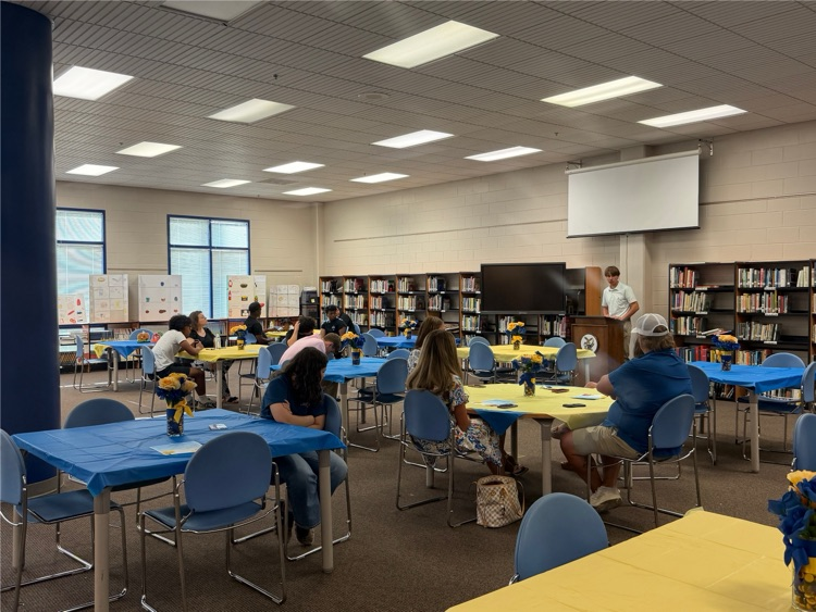 Library with tables that have blue and yellow table clothes with small floral arrangements. a male student is standing at the front talking to the group