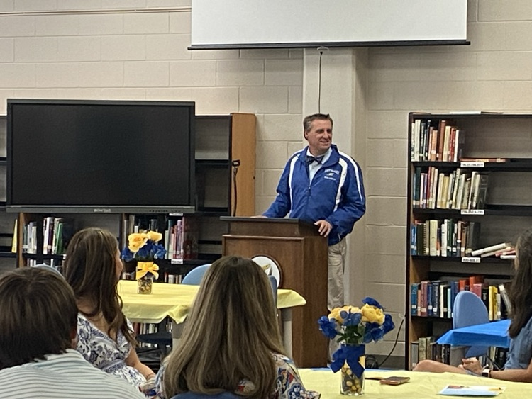 Library with tables that have blue and yellow table clothes with small floral arrangements. Dr. Carroll is standing at the front talking to the group