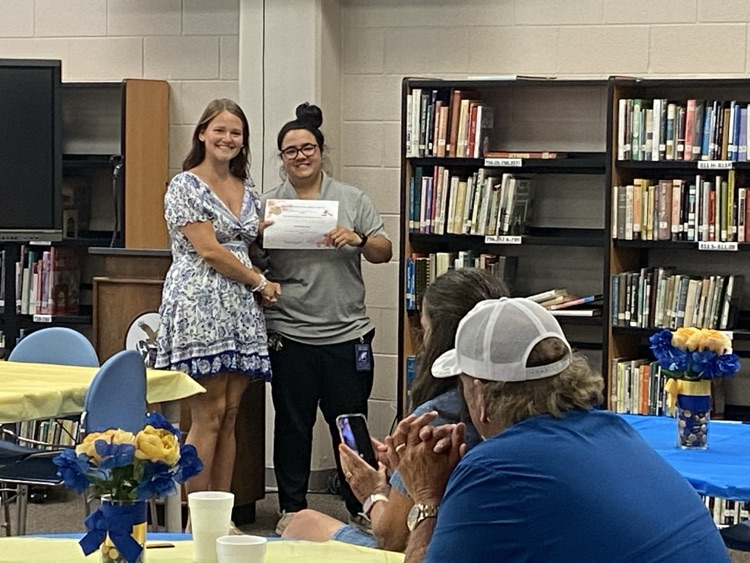 a student in a dress is presented a certificate by Ms. McMillian in black pants and a grey shirt.