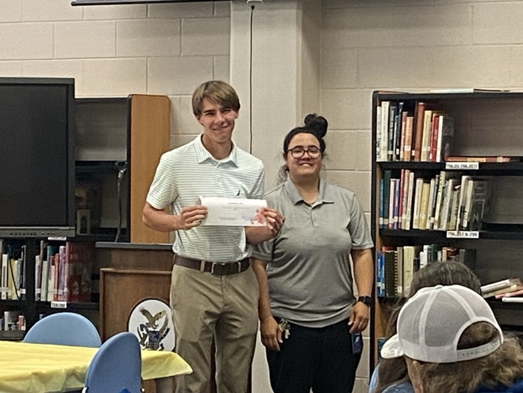 a student in a kaki pants and a white polo is presented a certificate by Ms. McMillian in black pants and a grey shirt.