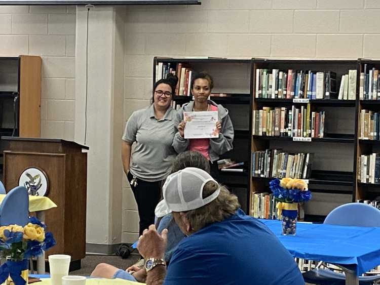 a student in a pink tshirt and grey jacket is presented a certificate by Ms. McMillian in black pants and a grey shirt.